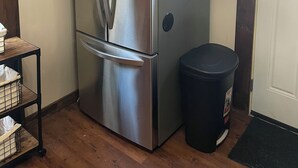 A stainless steel refrigerator with wooden upper cabinets, a trash can, and a small table with baskets.