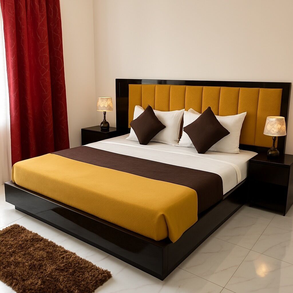 Uma cama bem arrumada com cabeceira amarela, duas mesas de cabeceira com abajures, uma cortina vermelha e um tapete marrom.