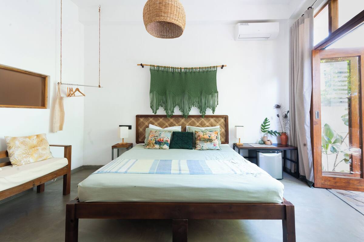 A bedroom with a bed, a hanging macrame wall hanging, a wooden headboard, a bedside table, and a view of greenery through a glass door.