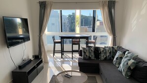 A modern living room with a flat-screen TV, a sofa with patterned cushions, a round coffee table, and a dining table with chairs. - 4-bedroom condo Steps from Ocean w/Pier, parking, WiFi, AC, parks & shopping. (Santo Domingo)