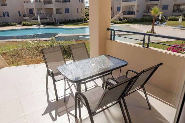 Un balcón con mesa y sillas de vidrio, con vista a una piscina y un área ajardinada con sillones y sombrillas.