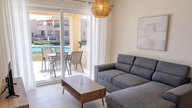 Un salon moderne avec un canapé gris, une table basse en bois et un téléviseur posé sur un meuble en bois. Une porte vitrée donne accès à un espace extérieur comprenant une piscine et des chaises.