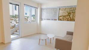 Un salon moderne avec un canapé beige, deux petites tables blanches et une grande fenêtre équipée de stores.