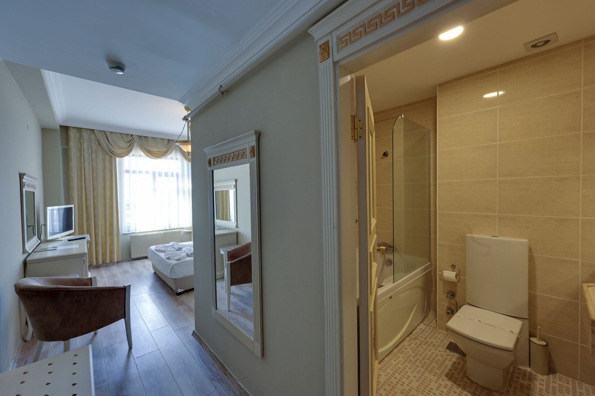 Ein Hotelzimmer mit einem Bett, einem Schreibtisch mit Computer, einem Sessel, einem großen Spiegel und einem Badezimmer mit Badewanne und Toilette.