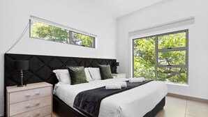 Un dormitorio moderno con cama, mesita de noche y dos ventanas con vistas a árboles.