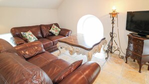 A living room with a brown leather sofa, a matching armchair, a coffee table, a TV on a wooden stand, and a floor lamp. - Crea The Mission Hall (Antrim)