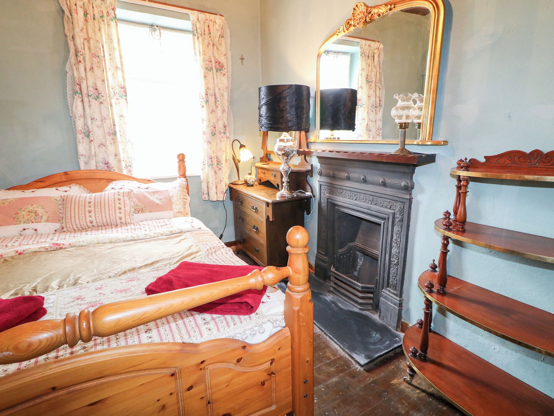 Une chambre à coucher avec un lit en bois, une commode, un miroir, une cheminée et une fenêtre ornée de rideaux à motifs floraux.