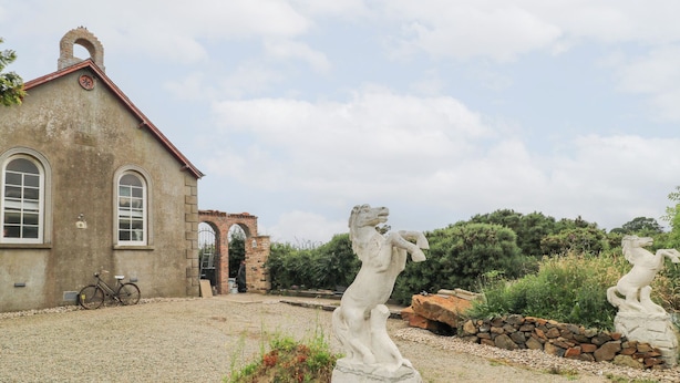 A building with a red roof and arched windows, a bicycle parked in front, and two white stone statues in a garden. - Crea The Mission Hall (Antrim)