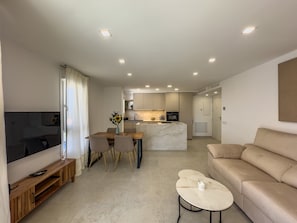 A modern living room with a TV, sofa, coffee table, and dining area with chairs and a vase of flowers.