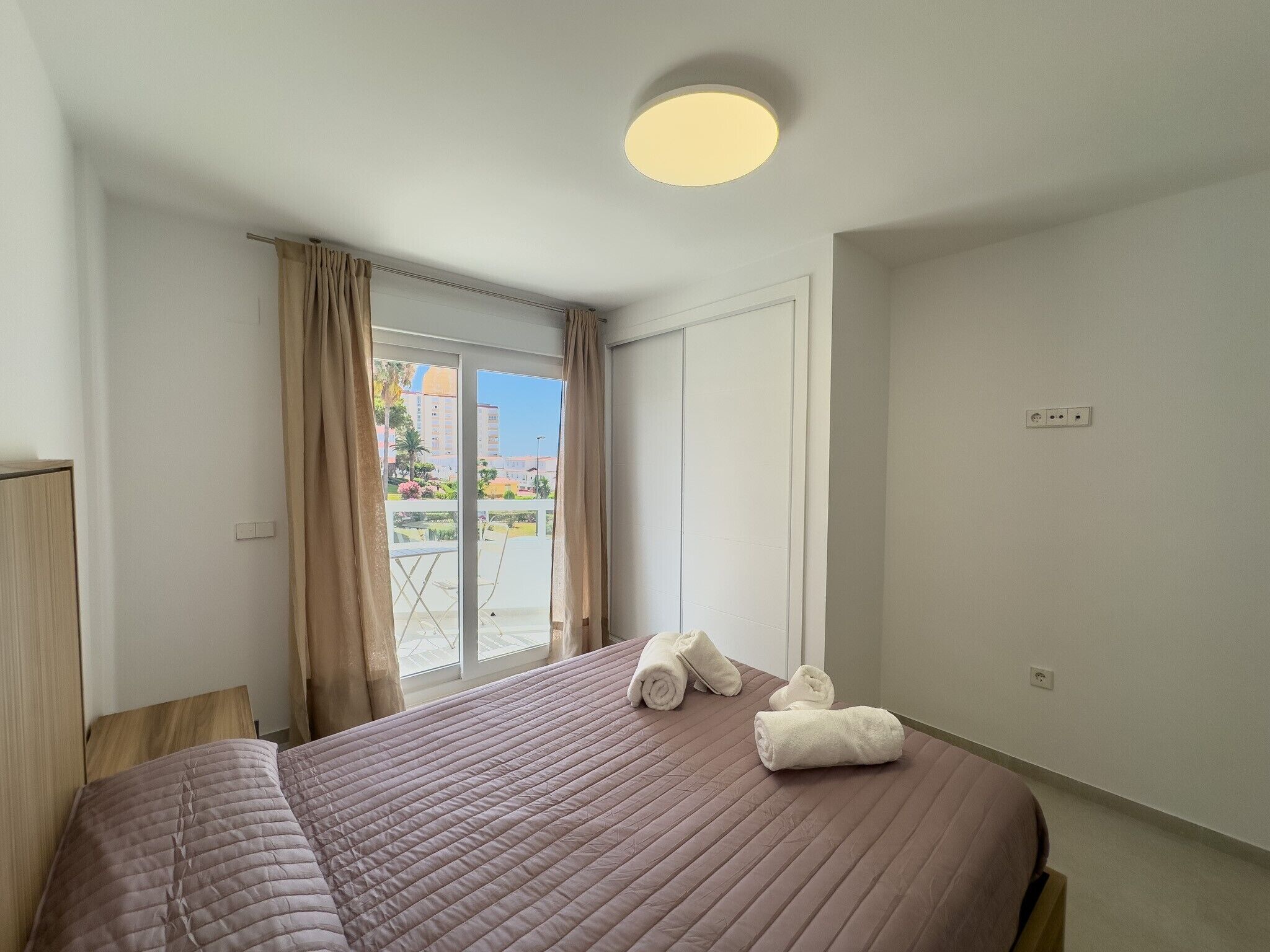 Un dormitorio con cama, ventana con cortinas, una lámpara de techo y vistas al exterior.