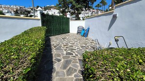 Un chemin de pierres mène à un bâtiment, avec une haie verte d’un côté et un mur blanc de l’autre.