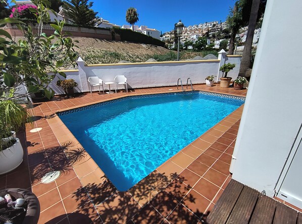 Une piscine rectangulaire ornée de motifs géométriques en mosaïque, entourée d’un mur blanc et offrant une vue sur une colline.