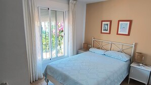 Une chambre à coucher avec un grand lit, une table de chevet, une fenêtre avec des rideaux et un mur orné de deux tableaux encadrés.