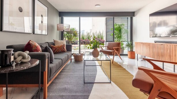 Un salon moderne avec un canapé, une table basse et des fauteuils, un téléviseur et de grandes fenêtres ornées de plantes.