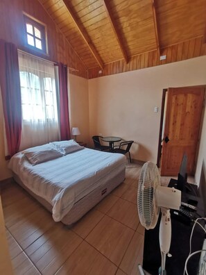 Una habitación con cama, mesa, silla, ventilador y puerta.