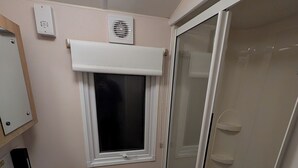 Ein kleines Badezimmer mit Waschbecken, einem Fenster mit Jalousie, einer Duschkabine mit Glastür und einem Wandlüfter.