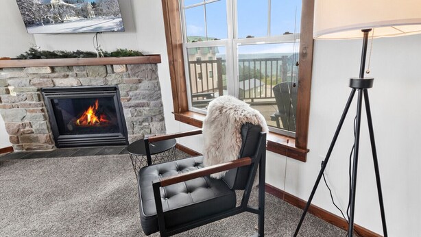 A cozy room with a fireplace, a comfortable chair, a floor lamp, and a large window offering a view of the outside. - Soaring Eagle Lodge #217 Beautiful upgraded Corner Unit Wraparound Balcony Ski-in/Ski-out Underground parking (Snowshoe)