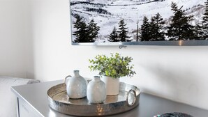 A modern living room with a flat-screen TV mounted on the wall, a grey sofa, a coffee table with decorative vases and a potted plant, and a remote control on the table. - Soaring Eagle Lodge #217 Beautiful upgraded Corner Unit Wraparound Balcony Ski-in/Ski-out Underground parking (Snowshoe)