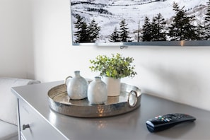 Una sala moderna con un televisor de pantalla plana montado en la pared, un sofá gris, una mesa de centro con jarrones decorativos y una planta en maceta, y un control remoto sobre la mesa.