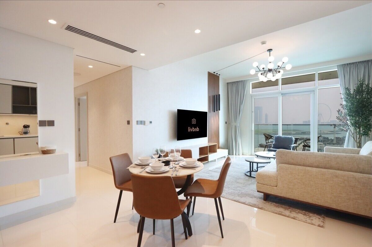 A modern living room with a dining area, a flat-screen TV mounted on the wall, and a large window offering a city view.