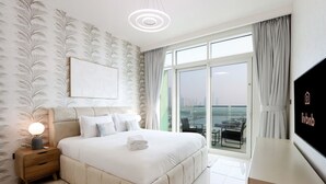 A modern hotel room with a large bed, a nightstand with a lamp, a view of the city through large windows, and a flat-screen TV on the wall.