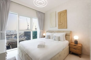 A hotel room with a large bed, a nightstand with a lamp, a view of a cityscape, and two abstract wall art pieces.