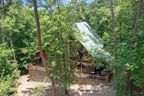 A cabin with a stone foundation, wooden deck, and a green roof, surrounded by dense forest.