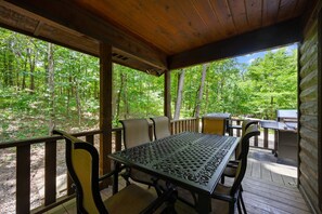 A covered outdoor seating area with a table and chairs, overlooking a forested area.