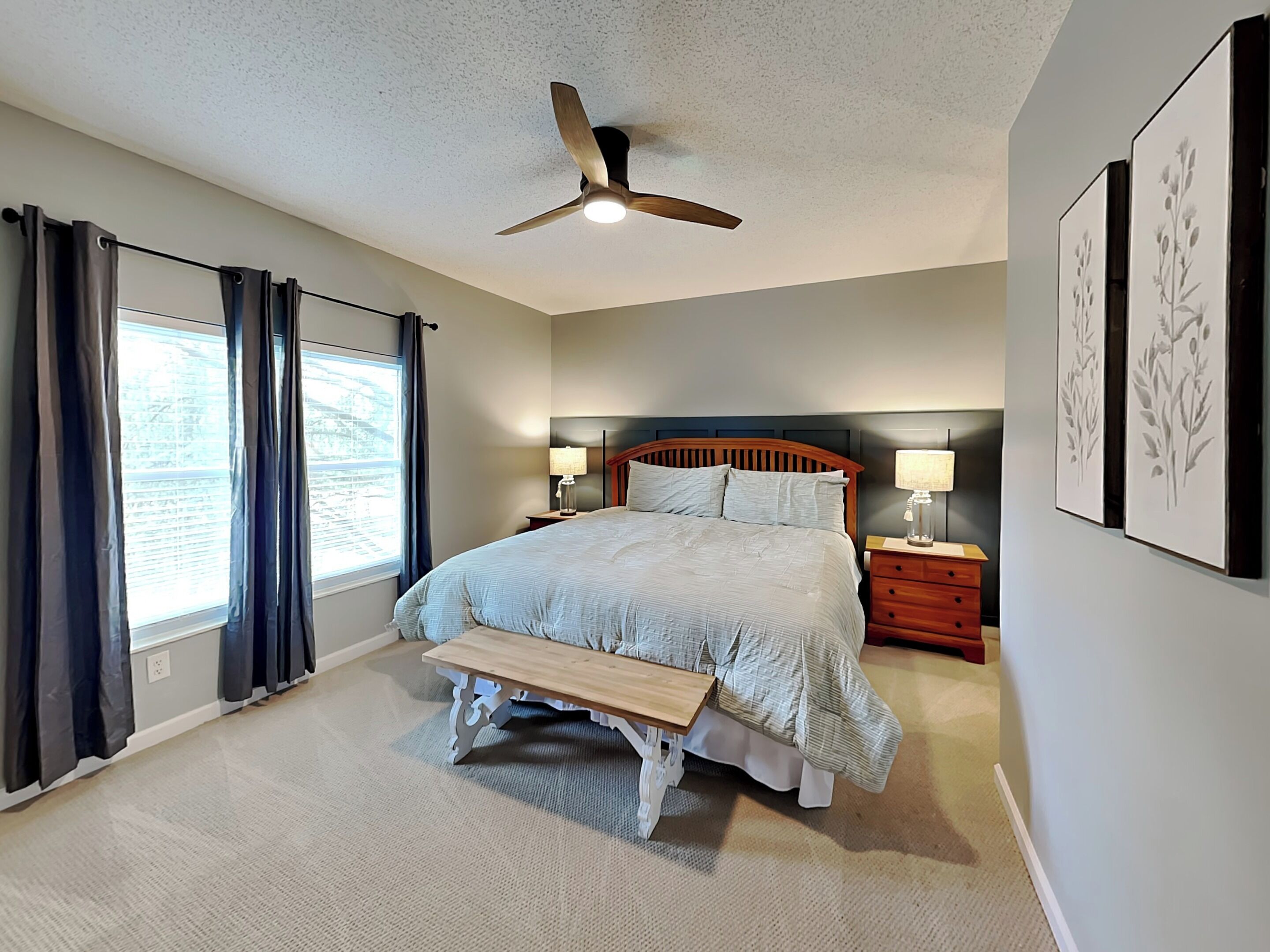 A bedroom with a bed, bedside tables, a bench, a ceiling fan, and artwork on the walls.
