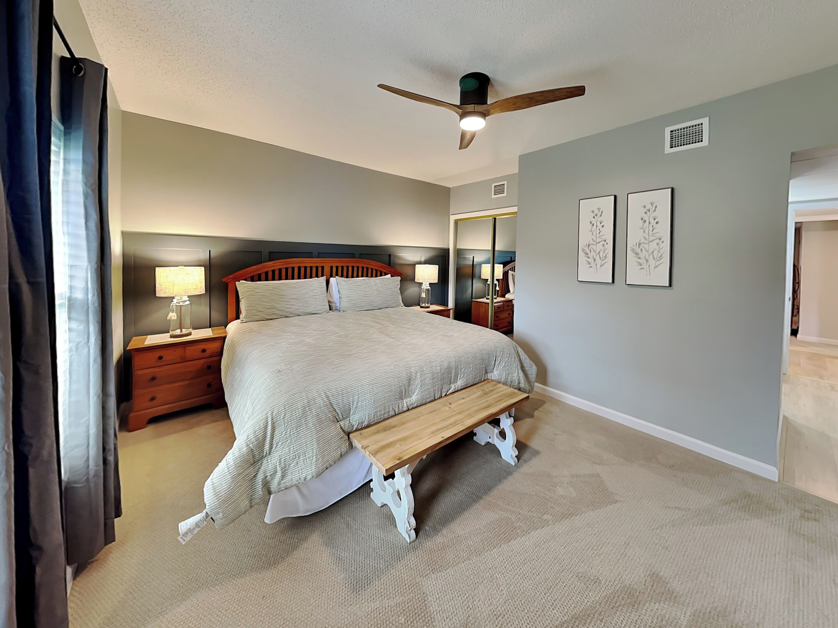 Une chambre à coucher comprenant un lit, des tables de chevet, un ventilateur de plafond, un banc et des œuvres d’art encadrées accrochées au mur.