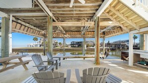 An outdoor patio with Adirondack chairs, a picnic table, and a grill area overlooking a body of water with a clear view of the beach and buildings in the background. - Reelview by Ryson Vacation Rentals (Galveston)