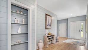 Een hal met lichtblauwe muren, houten vloeren en een witte deur aan het einde. Er staan decoratieve objecten op planken langs de muur, en boven de planken hangt een lijst met een afbeelding.