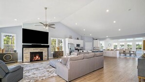 Een ruime woonkamer met een open haard, een grijze bank en een eethoek met uitzicht.
