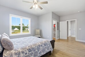 A bedroom with a large bed, a ceiling fan, a window with a view of a house, and a hallway leading to a closet.