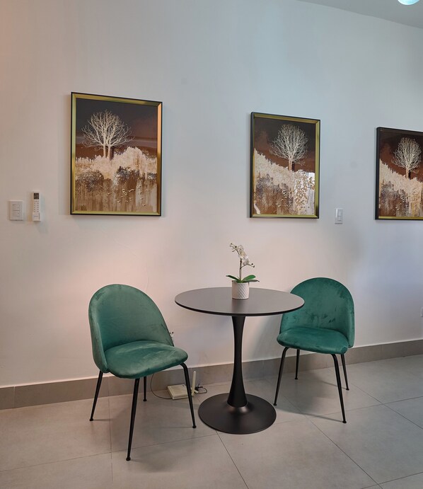A room with two green chairs around a small round table, a potted orchid, and three framed paintings on the wall.