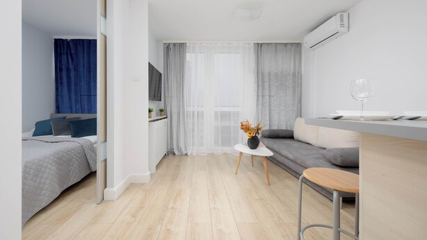 A modern, well-lit room with a bed, a sofa, a small table with a vase, and a bar area with a stool. - Comfortable Apartment in the Center | View of Skyscrapers, Air Conditioning (Warszawa)