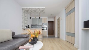 A modern living room with a grey sofa, a white coffee table with a magazine, and a vase with flowers. A kitchen is visible in the background with a microwave, oven, and cabinets. - Comfortable Apartment in the Center | View of Skyscrapers, Air Conditioning (Warszawa)