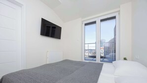 A bedroom with a bed, a TV mounted on the wall, a window with a view of buildings, and a radiator. - Functional Apartment for 6 Persons | Air Conditioning, Balcony (Warszawa)