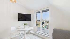 Una habitación moderna y minimalista con una mesa redonda de comedor, sillas blancas, un televisor de pantalla plana montado en la pared y un ventanal con vistas a la ciudad.