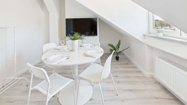 A modern dining area with a round table set for four, white chairs, a potted plant, and a window with a view of buildings.