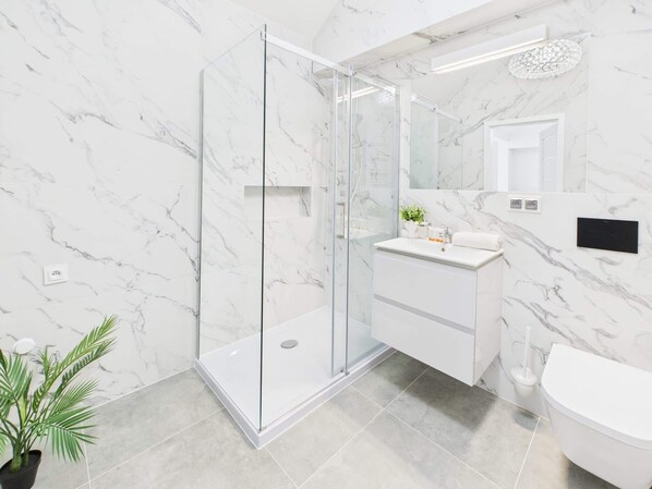 A modern bathroom with a glass shower enclosure, white marble walls, a white sink, and a toilet.