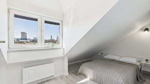 Ein Schlafzimmer mit schrägverdeckter Decke, zwei Fenstern mit Jalousien, einem Heizkörper, einem Bett mit grauer Bettdecke und einem kleinen Nachttisch mit Lampe.