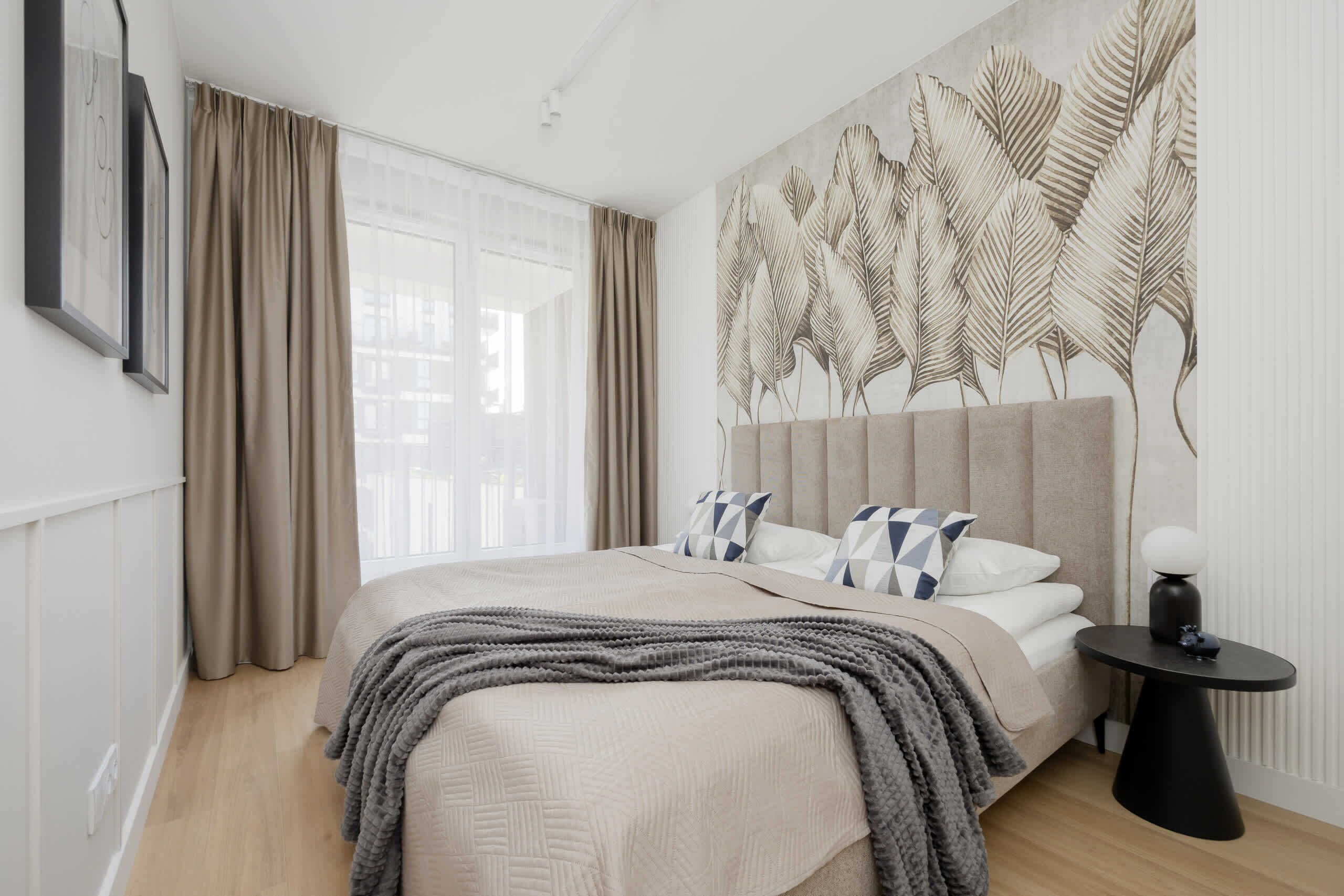 Un dormitorio moderno con una cama grande, cortinas color beige, un mural en la pared y una mesita de noche pequeña.