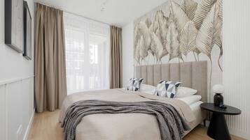 Un dormitorio moderno con una cama grande, cortinas color beige, un mural en la pared y una mesita de noche pequeña.