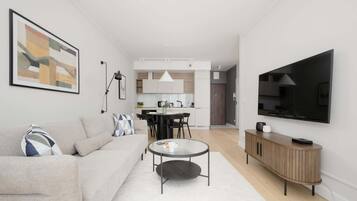 Una sala de estar moderna con sofá, mesa de centro y televisor. Se observa una cocina al fondo con zona de comedor.