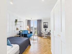 A modern hotel room with a large bed, a blue sofa, a dining table with chairs, and a city view through the window.