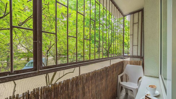 Un balcón con vista a árboles verdes, una mesa blanca con un plato de comida y una taza, y una silla blanca.