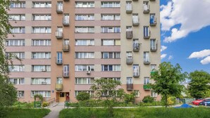 Un edificio residencial de varios pisos con balcones, una entrada central y un estacionamiento con coches.