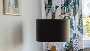 Una habitación con un sofá color mostaza, una lámpara de pie con pantalla negra y cortinas con estampado de hojas tropicales.