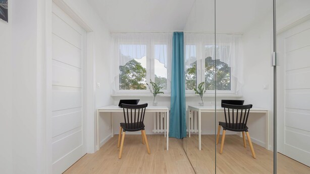 Una habitación con dos escritorios y sillas, una ventana con vista a árboles y una cortina color turquesa.
